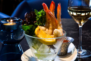 A shrimp cocktail served in a martini glass with cocktail sauce, lemon, and ice, accompanied by a glass of white wine.