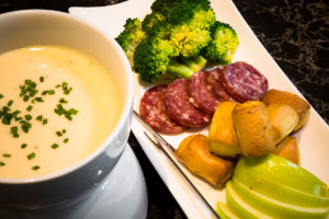 A cheese fondue pot with melted cheese, served with bread, apples, broccoli, and salami.