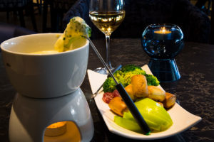 A cheese fondue pot with melted cheese, served with bread, apples, broccoli, and salami.