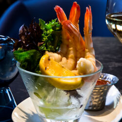 A shrimp cocktail served in a martini glass with cocktail sauce, lemon, and ice, accompanied by a glass of white wine.