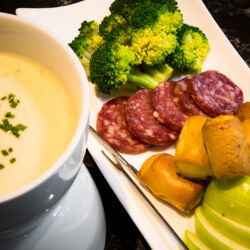 A cheese fondue pot with melted cheese, served with bread, apples, broccoli, and salami.