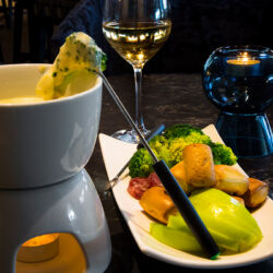 A cheese fondue pot with melted cheese, served with bread, apples, broccoli, and salami.